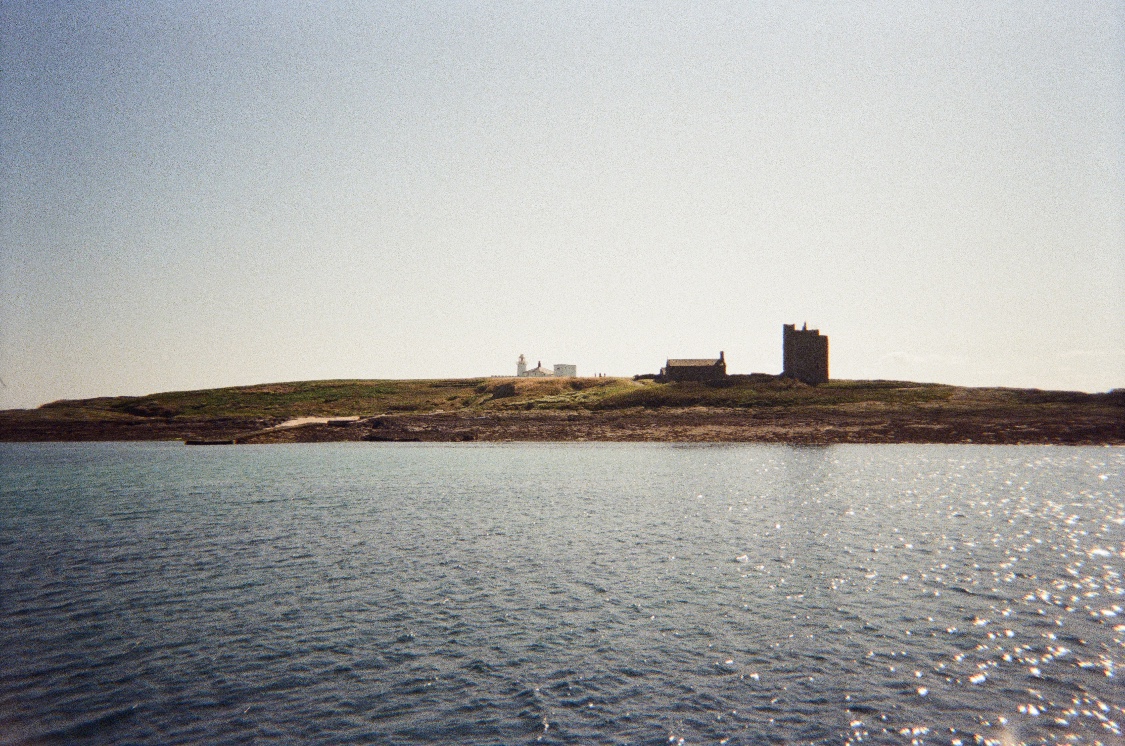A low green island seen across sparkling water, with a small white lighthouse and a ruined stone tower on the horizon