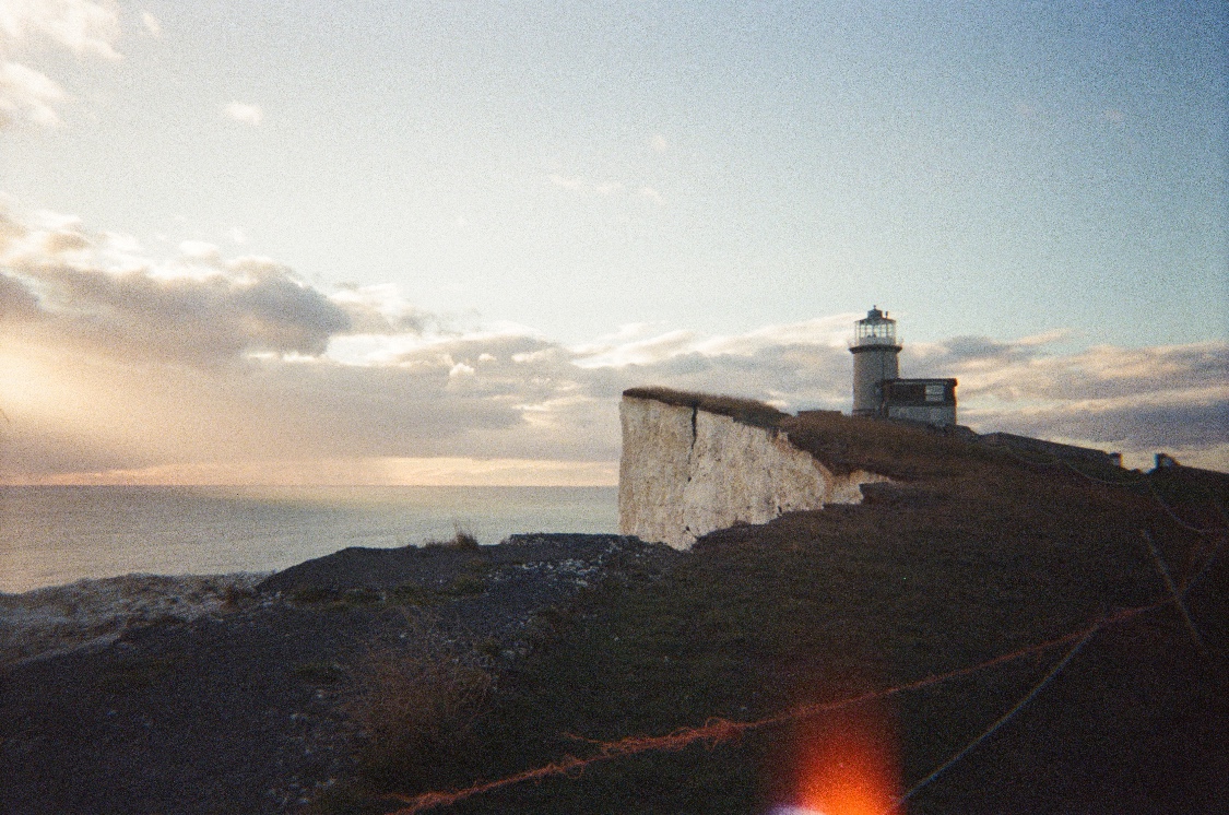 A lighthouse on top of a white chalk cliff at dusk, the sea and a low sun behind clouds, with a red light leak in the corner