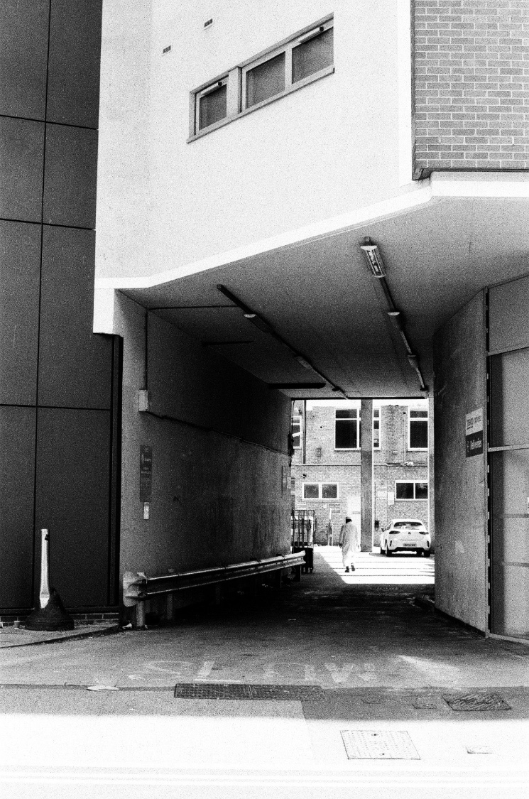 Black and white photo of a shaded passage under a building, opening onto a bright courtyard where a figure in white walks away
