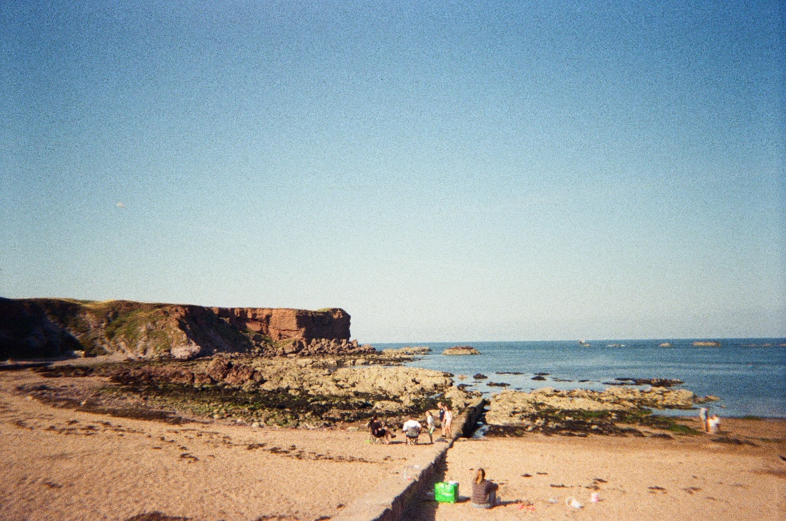 A sandy beach at low tide with rock pools, red sandstone cliffs and a few people sitting on the sand under a clear blue sky