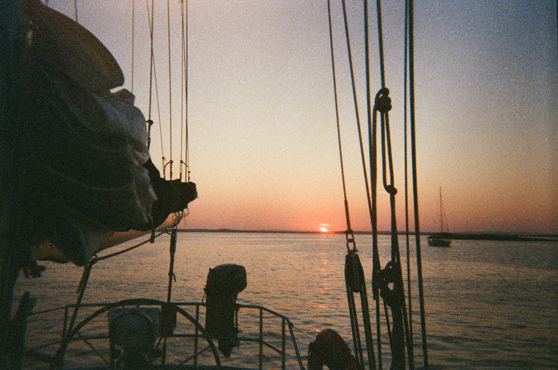 The sun setting over calm water, seen from the deck of a sailing boat between its rigging, with another boat moored in the distance