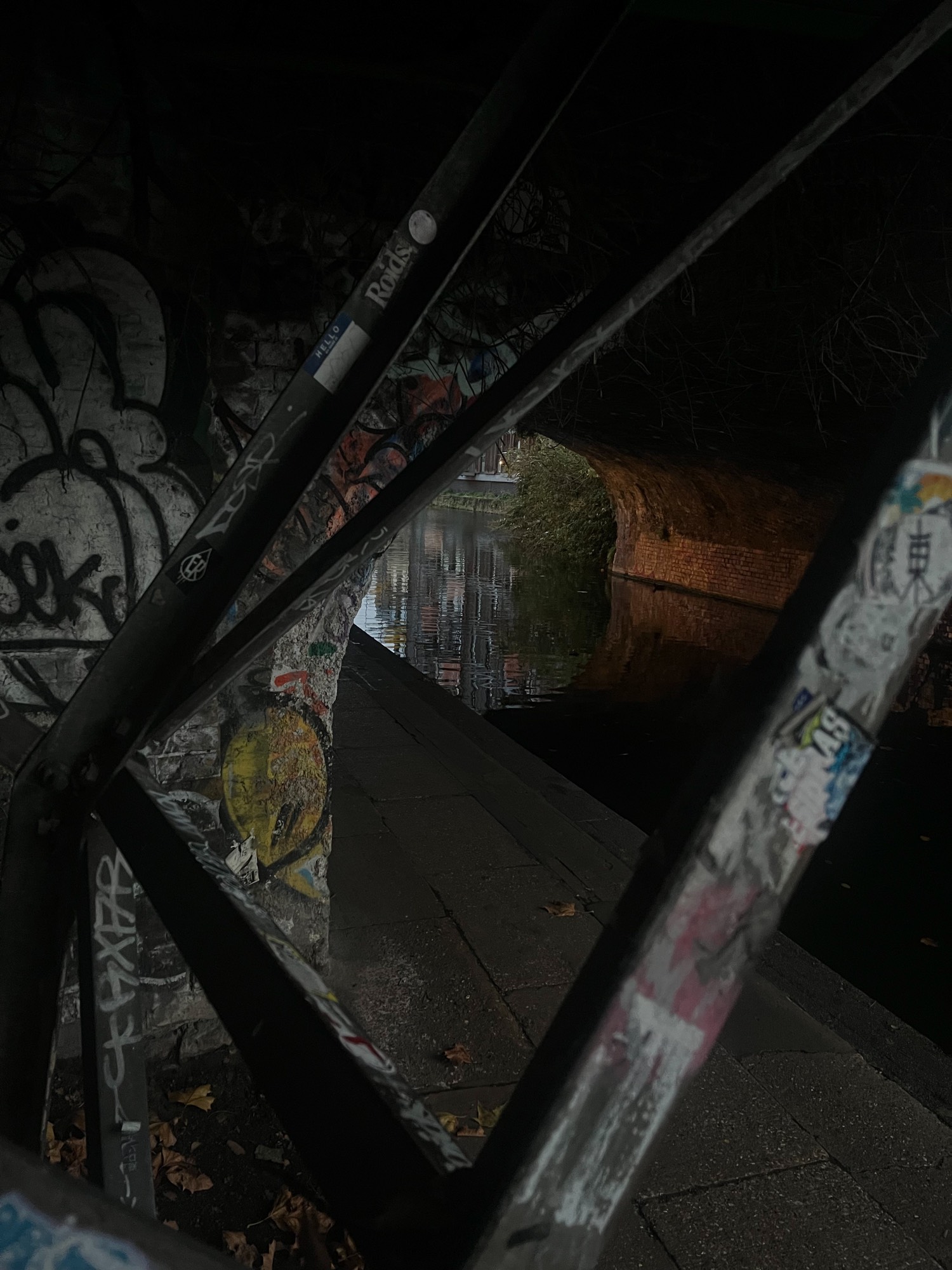 Canal towpath seen through sticker-covered railings, with warm light on the brick arch of a bridge